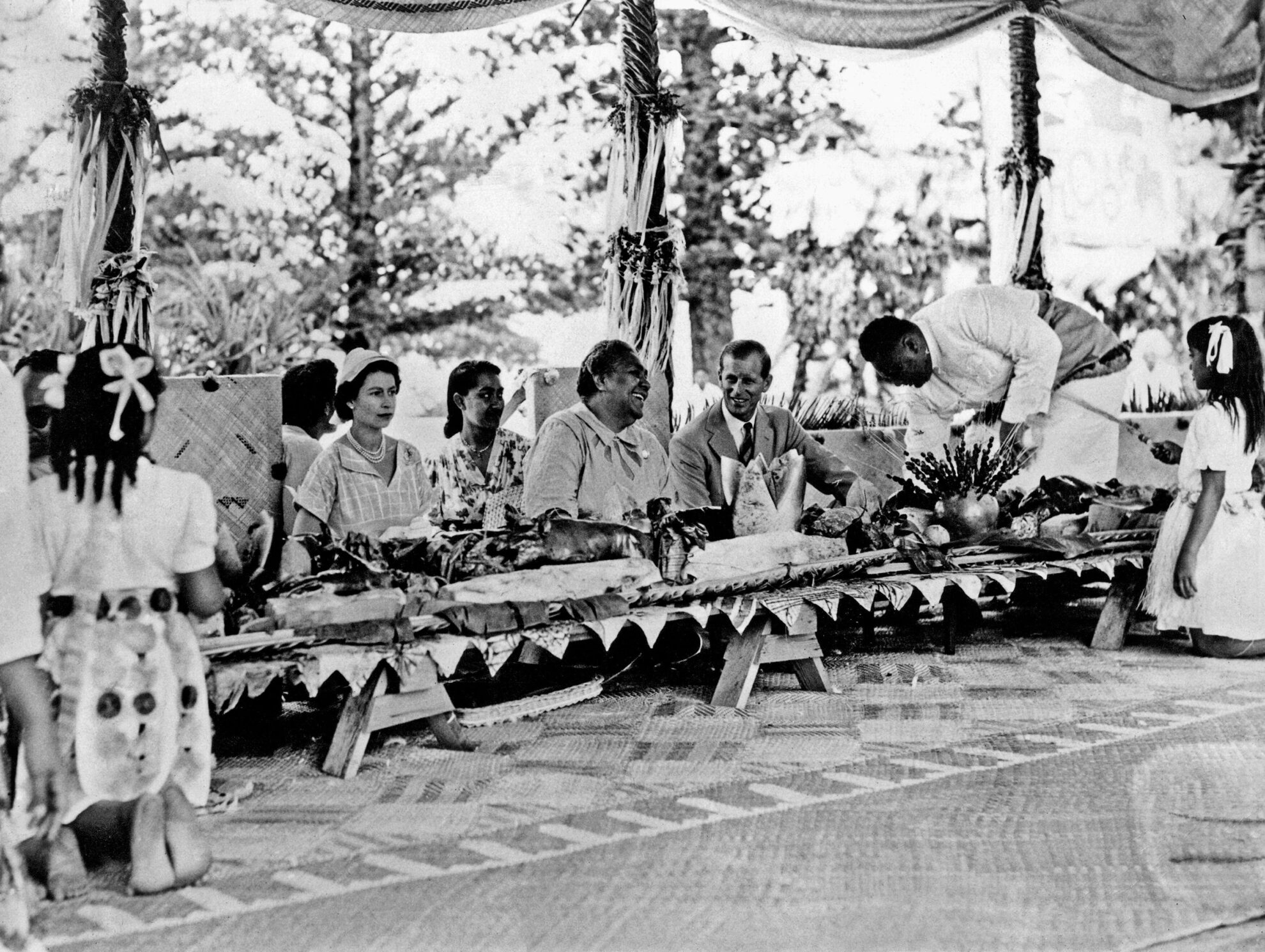 Ratu Elizabeth II dan Pangeran Philip dengan Ratu Tonga di sebuah pesta di mana babi panggang ditempatkan di depan setiap orang, selama Tur Kerajaan Persemakmuran 19-20 Desember 1953.