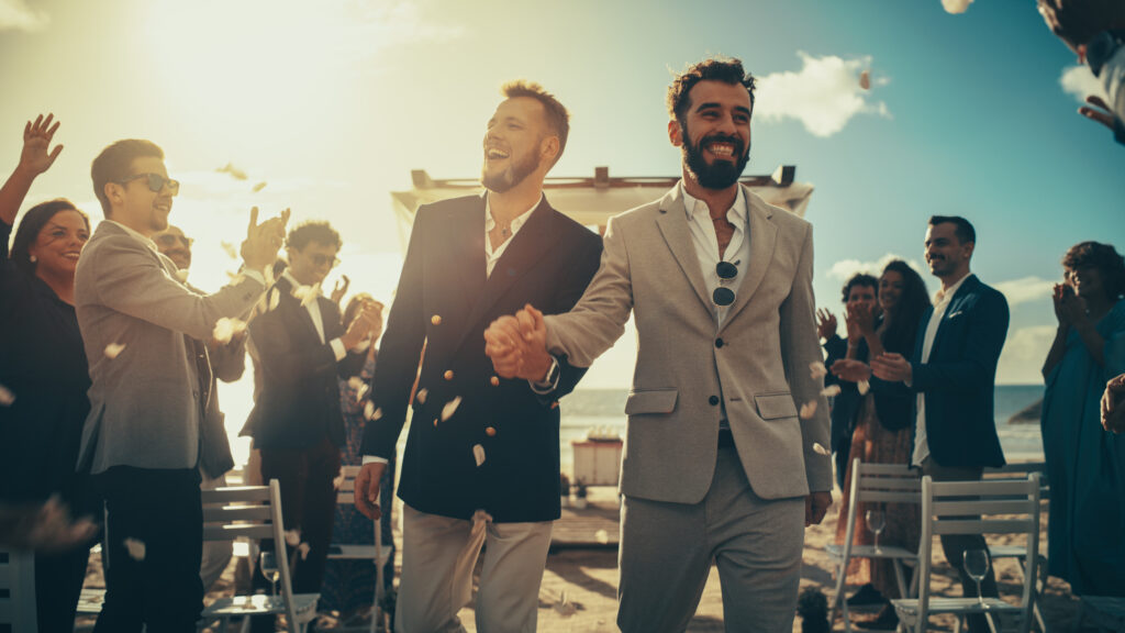 Handsome Gay Couple Walking Up the Aisle at Outdoors Wedding Ceremony Venue Near Sea. Two Happy Men in Love Share Their Big Day with Diverse Multiethnic Friends. Authentic LGBTQ Relationship Goals.